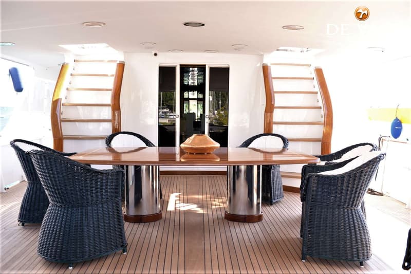 Een houten dek met een tafel en zes stoelen, geflankeerd door twee trappen en een glazen schuifdeur.