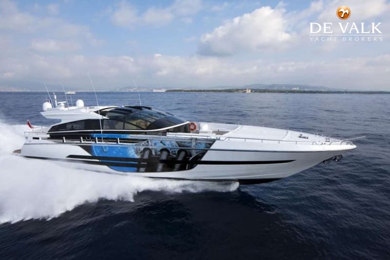 Een witte motorjacht met blauwe en zwarte accenten vaart op open water, met een kustlijn op de achtergrond.