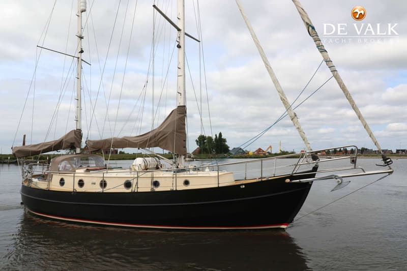Een zwarte zeilboot met een crèmekleurige opbouw en bruine zeilhoezen ligt in het water onder een bewolkte lucht.