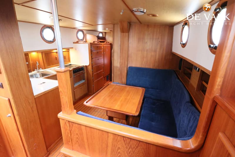De foto toont een houten interieur van een boot met een kombuis, een zithoek met blauwe kussens en een tafel.
