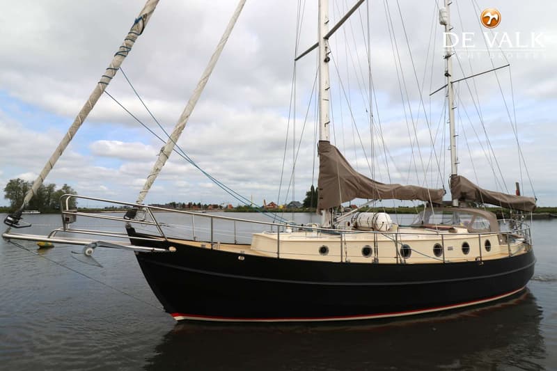 Een donkerblauwe zeilboot met een crème opbouw en een bruine zeilhuik ligt in het water onder een bewolkte hemel.