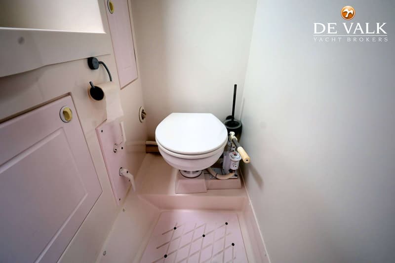 Een witte scheepstoilet met een handpomp en een toiletborstel in een kleine, lichte ruimte.