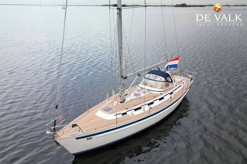 Een witte zeilboot met een houten dek en blauwe accenten ligt op rustig water, met een Nederlandse vlag aan de achtersteven.