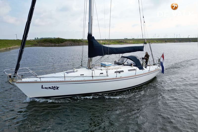 Een witte zeilboot met de naam "Loenatik" vaart op water, met een persoon aan het roer en een Nederlandse vlag achterop.