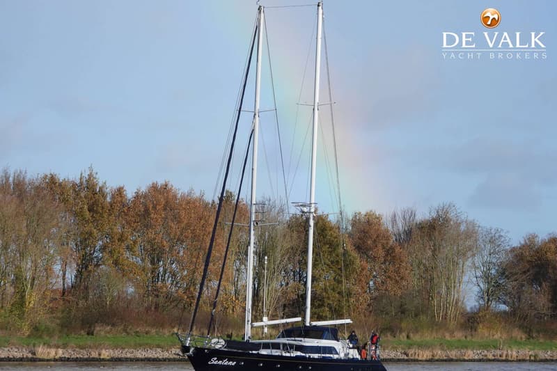 Een donkerblauwe zeilboot met twee masten en de naam "Santana" vaart op het water, met een regenboog op de achtergrond.