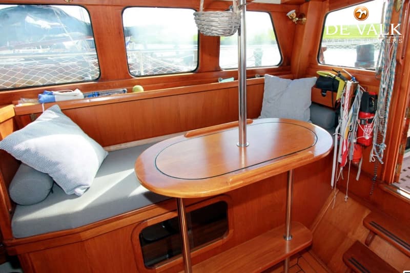 Een houten interieur met een tafel, banken met kussens, ramen en diverse scheepsbenodigdheden.