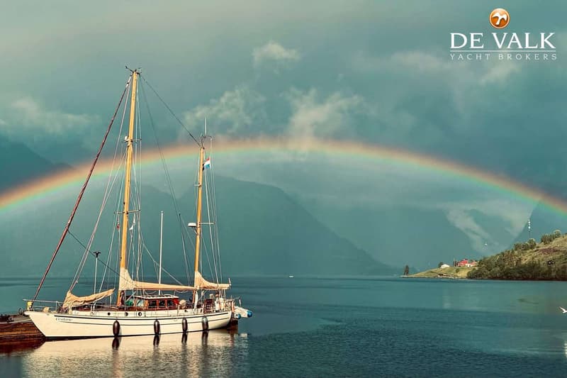 Een witte zeilboot met twee masten en een Nederlandse vlag ligt in een fjord met een regenboog op de achtergrond.