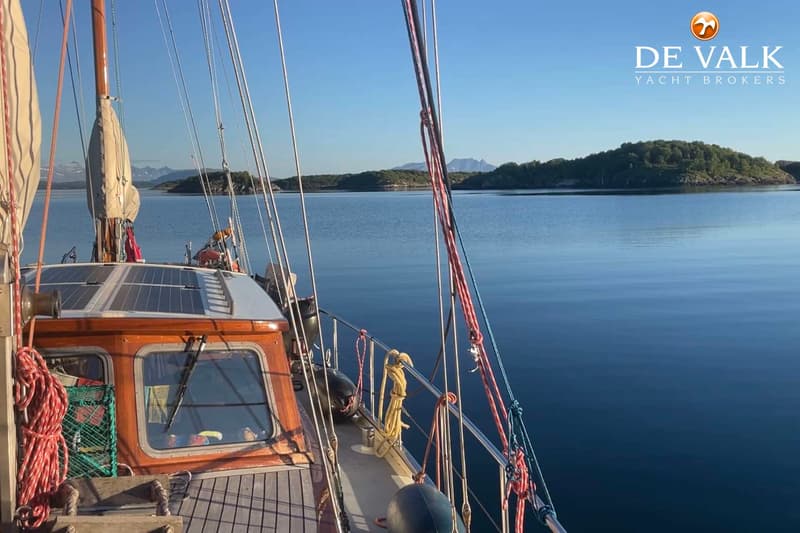 Een zeilboot met een houten kajuit en zonnepanelen op het dak vaart op rustig water met eilanden en bergen op de achtergrond.