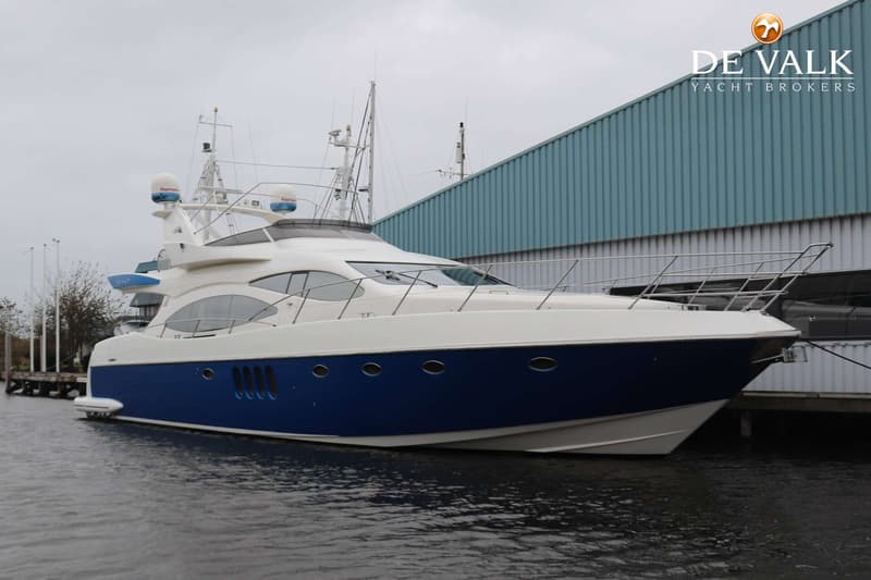 Een grote, witte en blauwe motorboot ligt aangemeerd aan een steiger, met een gebouw op de achtergrond. — Azimut 68 Plus