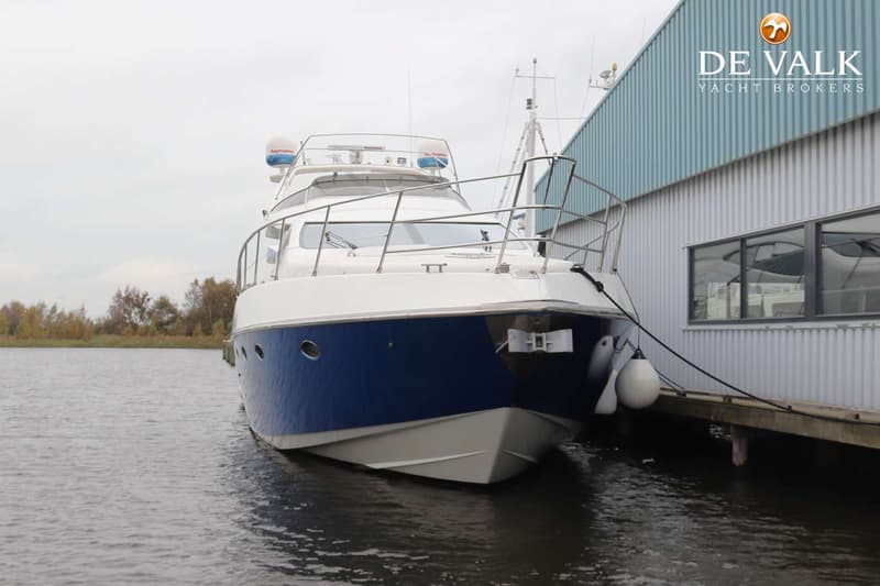 Een witte en blauwe motorboot ligt afgemeerd aan een steiger naast een gebouw met een logo van "DE VALK YACHT BROKERS".