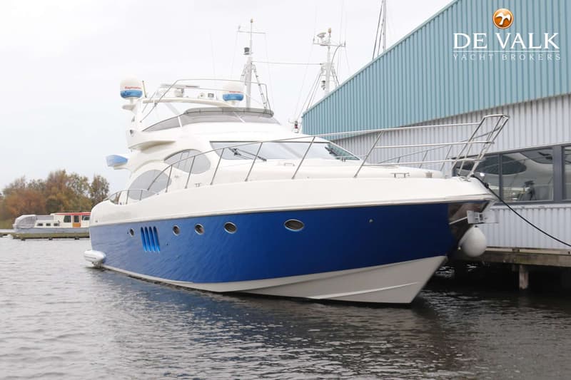 Een witte motorboot met een blauwe romp ligt afgemeerd aan een steiger naast een lichtblauwe loods. — Azimut 68 Plus