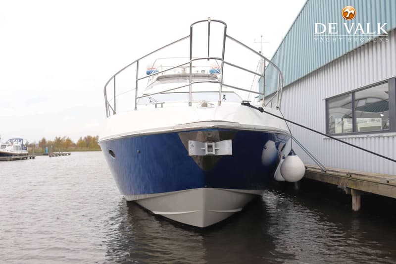 Een witte en blauwe boot ligt afgemeerd aan een houten steiger naast een gebouw met de tekst "DE VALK YACHT BROKERS".