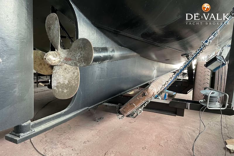 Een close-up van de onderkant van een zwarte boot met een driebladige schroef en een anker, ondersteund door stalen balken.