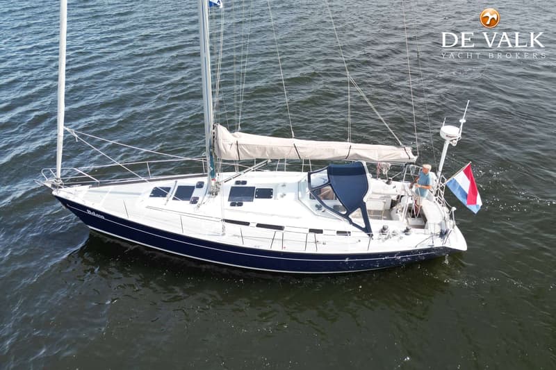 Een zeilboot met een donkerblauwe romp en witte opbouw vaart op water, met een man aan het roer en een Nederlandse vlag.