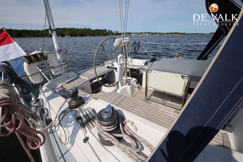 Een deel van het dek van een zeilboot met een stuurwiel, lieren, touwen en een Nederlandse vlag, met water en land op de…