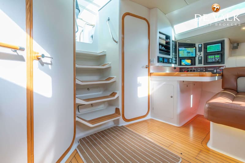 De foto toont het interieur van een boot met een trap, een deur, een navigatiehoek en een bruine leren bank.