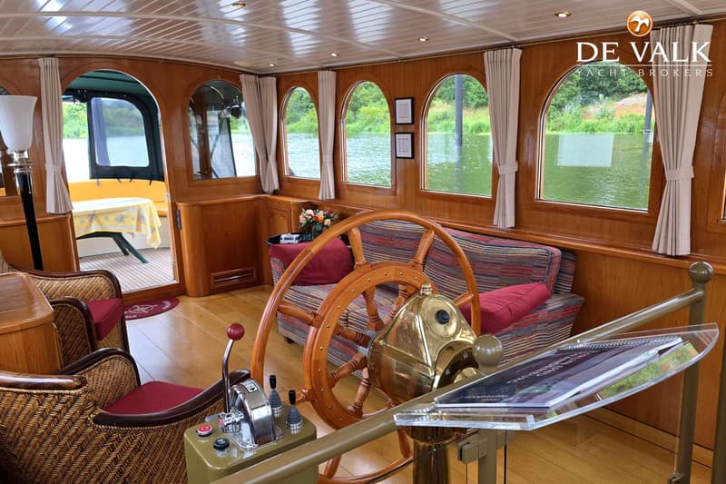 Een houten interieur van een boot met een stuurwiel, bank, stoelen en ramen die uitkijken op water en groen.