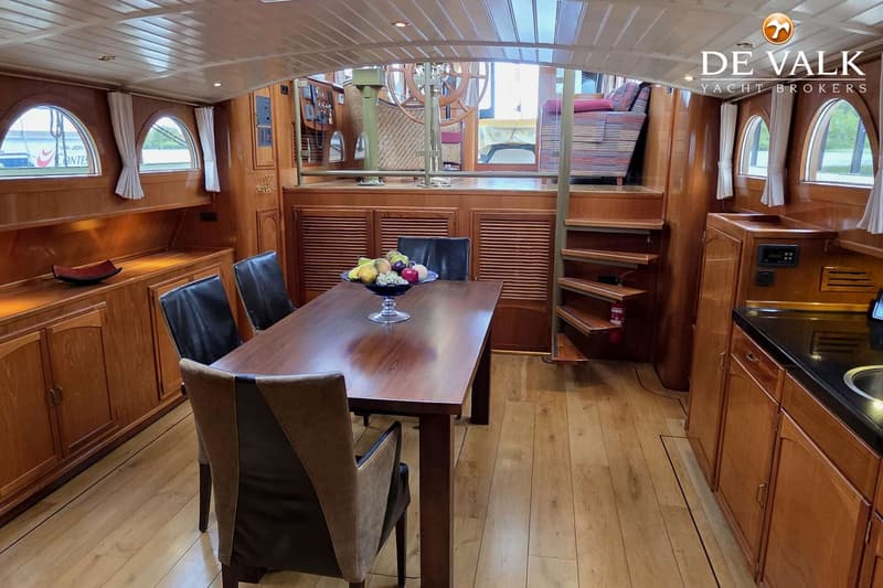 Een houten interieur van een boot met een eettafel, vier stoelen, een fruitschaal en een trap naar een hoger dek.