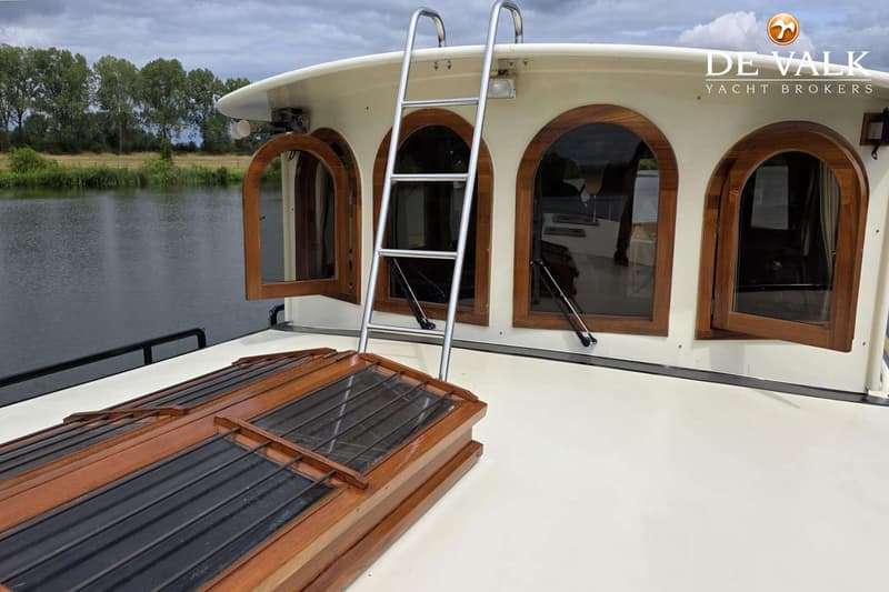 Een boot met een wit dek, houten ramen en een metalen ladder, varend op een rivier met bomen op de achtergrond.