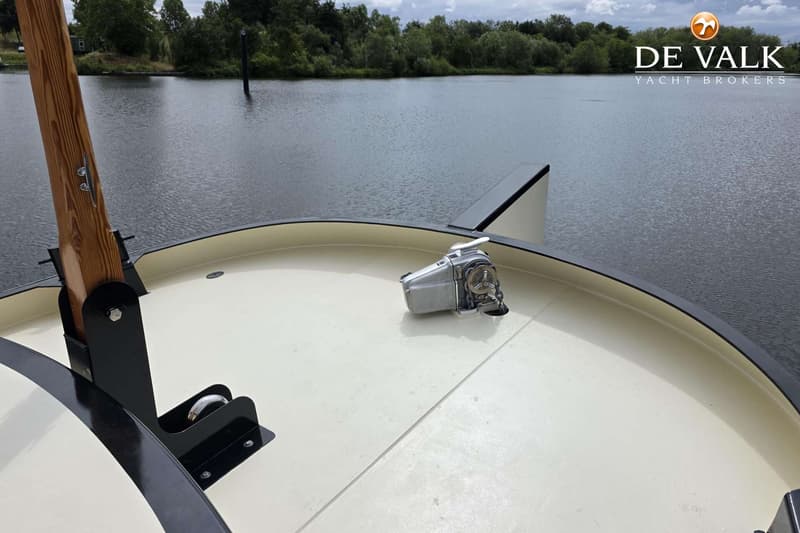 De foto toont het voordek van een boot met een ankerlier en een houten mast, met water en bomen op de achtergrond.