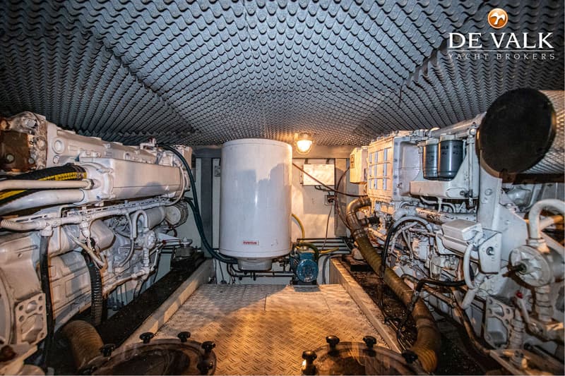 De foto toont een machinekamer met twee grote witte motoren, een boiler en een geribbeld plafond.