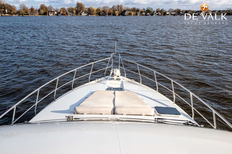 De foto toont de boeg van een wit jacht met reling, twee ligkussens en een ankerlier, varend op water met huizen op de…