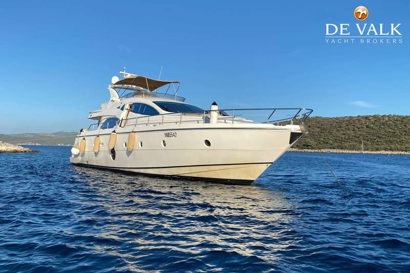 Een witte motorjacht met beige stootwillen en een beige bimini-top ligt op blauw water, met een heuvelachtige kustlijn op…