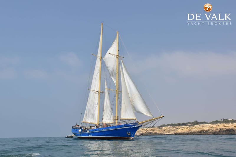 Een blauwe tweemaster zeilboot met witte zeilen vaart op open water, met een rotsachtige kustlijn op de achtergrond.