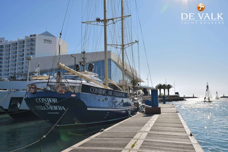 Een donkerblauwe zeilboot met twee masten ligt aangemeerd aan een houten steiger, met een hotel op de achtergrond.