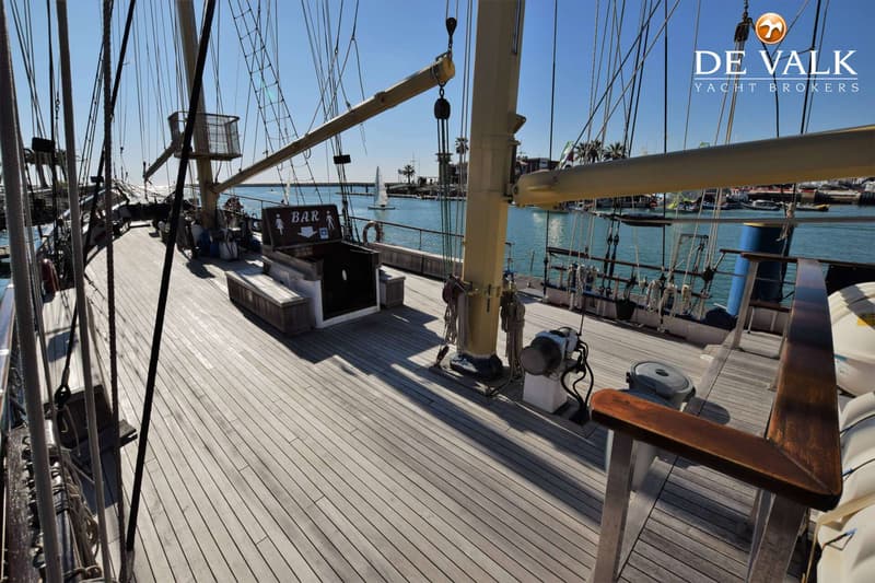 Een houten dek van een zeilschip met een bar, masten en touwen, met op de achtergrond een haven en de zee.