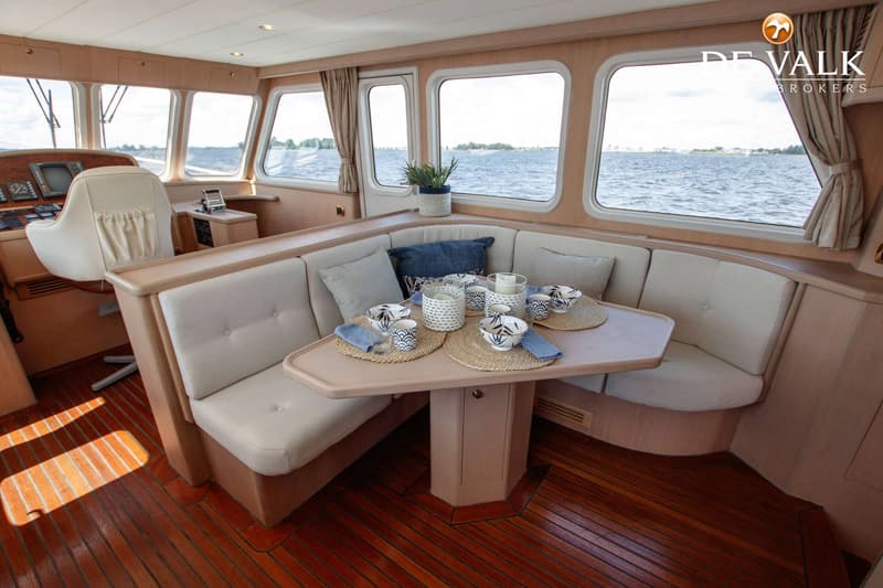 Een interieur van een boot met een houten vloer, een beige bank met kussens, een tafel met servies en ramen met uitzicht op…