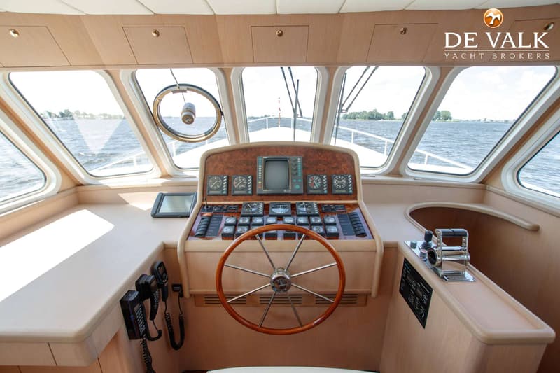 De foto toont het stuurhuis van een boot met een houten stuurwiel, diverse meters en schermen, en uitzicht op water.