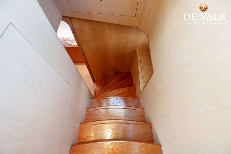 Een houten trap met vijf treden leidt naar beneden, met een bed zichtbaar aan de linkerkant en een kastje aan de rechterkant.