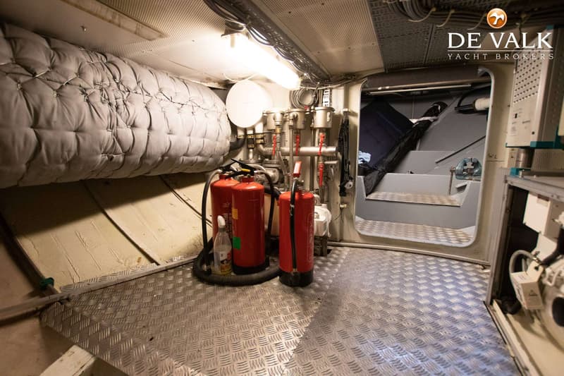 Een machinekamer met twee rode brandblussers, een geïsoleerde pijp en een trap naar een hoger dek.