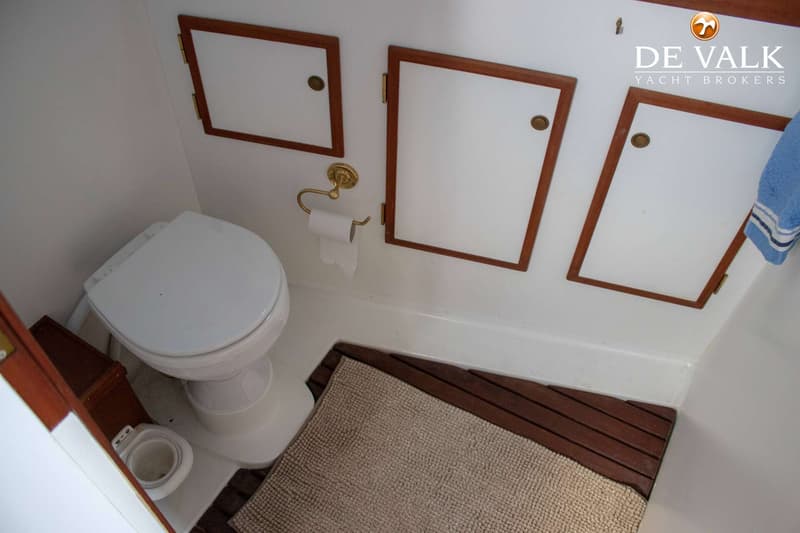 Een witte toiletruimte met een toilet, een toiletrolhouder, drie opbergkastjes en een houten vloerrooster met een badmat.