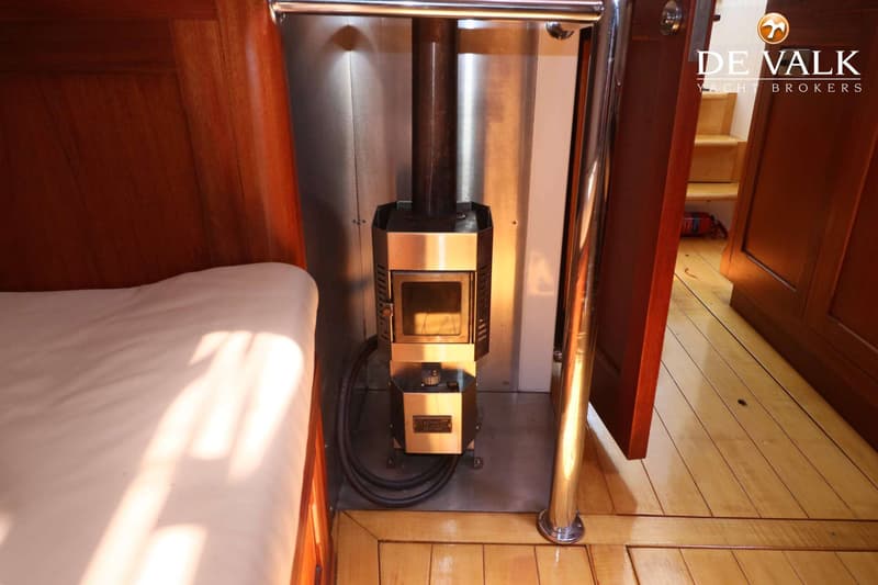 Een metalen kachel met een schoorsteenpijp staat op een houten vloer naast een bed met een wit matras.