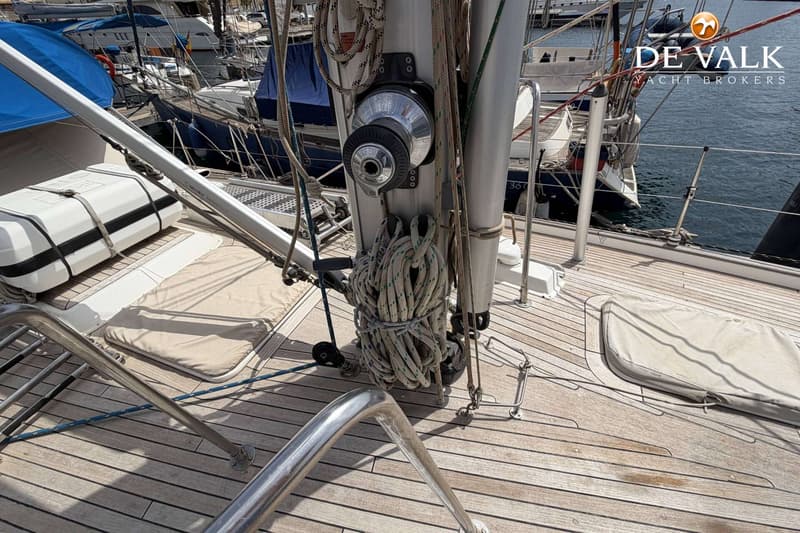 Een dek van een zeilboot met een lier, touwen, een reddingsvlot en een reling, met andere boten op de achtergrond.