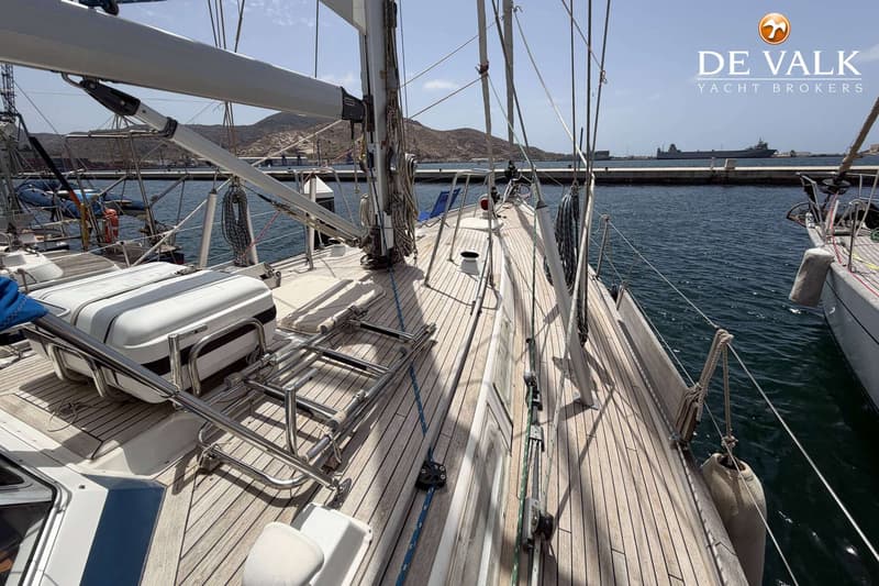Een zeilboot met een teakhouten dek ligt in een haven met bergen op de achtergrond. — Hallberg Rassy 46