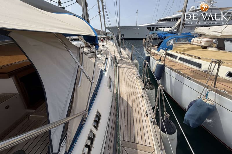 Twee zeilboten liggen afgemeerd naast elkaar, met een groot jacht op de achtergrond en een logo van "DE VALK YACHT BROKERS".