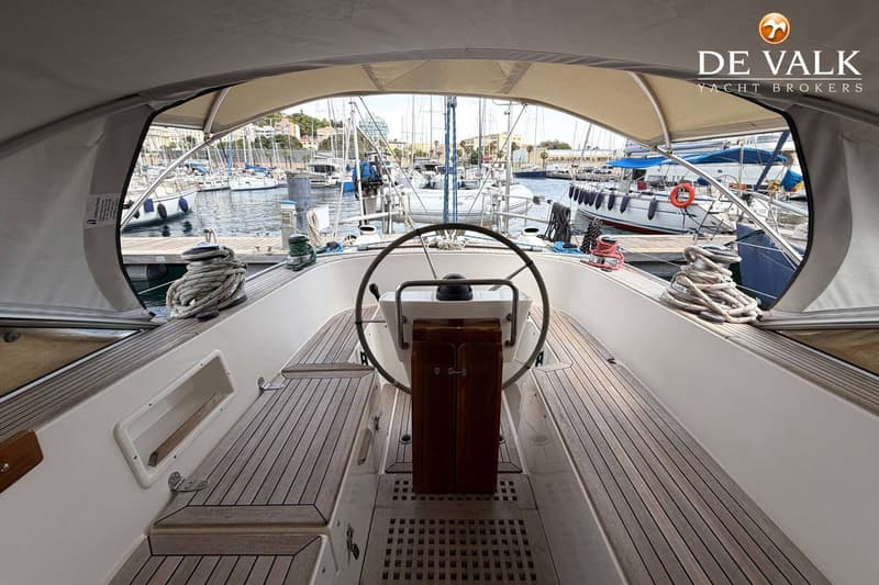 De foto toont het dek van een zeilboot met een stuurwiel, omringd door teakhouten vloeren en een uitzicht op een jachthaven.