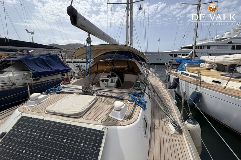 Een zeilboot met teakhouten dek, zonnepanelen en een stuurwiel in de kuip, afgemeerd tussen andere boten. — Hallberg Rassy 46