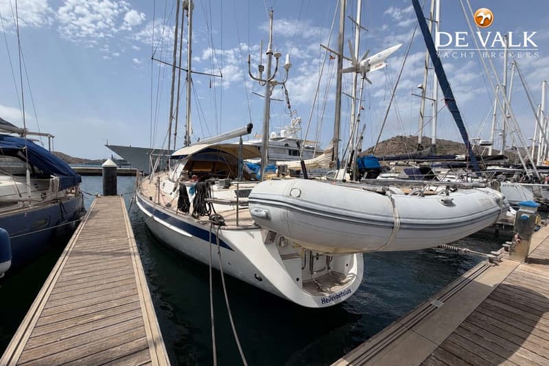 Een witte zeilboot met blauwe strepen en een bijboot hangend aan de achtersteven, ligt aangemeerd aan een houten steiger.