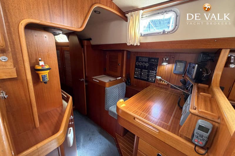 Een houten interieur van een boot met een navigatiehoek, een bankje en een patrijspoort met gordijn. — Hallberg Rassy 46