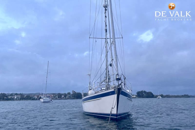 Een zeilboot met een witte romp en blauwe strepen ligt voor anker op het water, met een bewolkte lucht erboven.