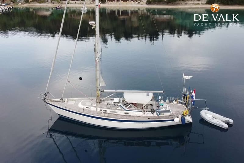 Een witte zeilboot met een blauwe streep ligt in rustig water, met een bijboot ernaast en een kustlijn op de achtergrond.