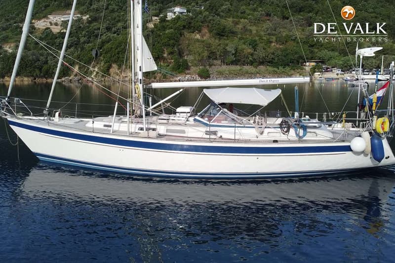 Een witte zeilboot met blauwe strepen ligt in het water met een groene, beboste kustlijn op de achtergrond.