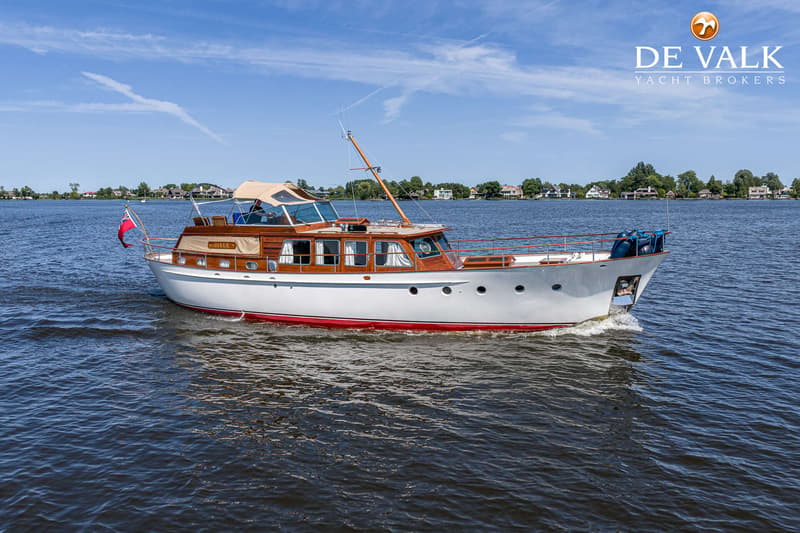 Een witte motorboot met houten opbouw en een rode waterlijn vaart over rustig water onder een blauwe lucht.