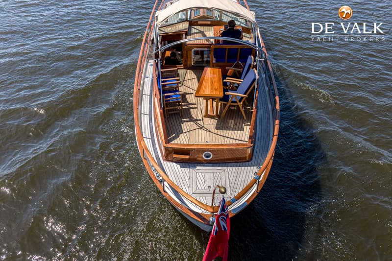 Een bovenaanzicht van een houten boot met een beige zonnescherm, varend op water, met een vlag aan de achtersteven.
