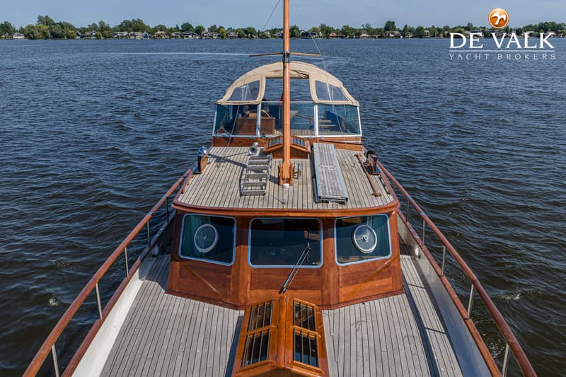 Een houten boot met een beige kap en een mast vaart op een waterweg met huizen langs de oever. — Kok Spitsgatkotter 16.50
