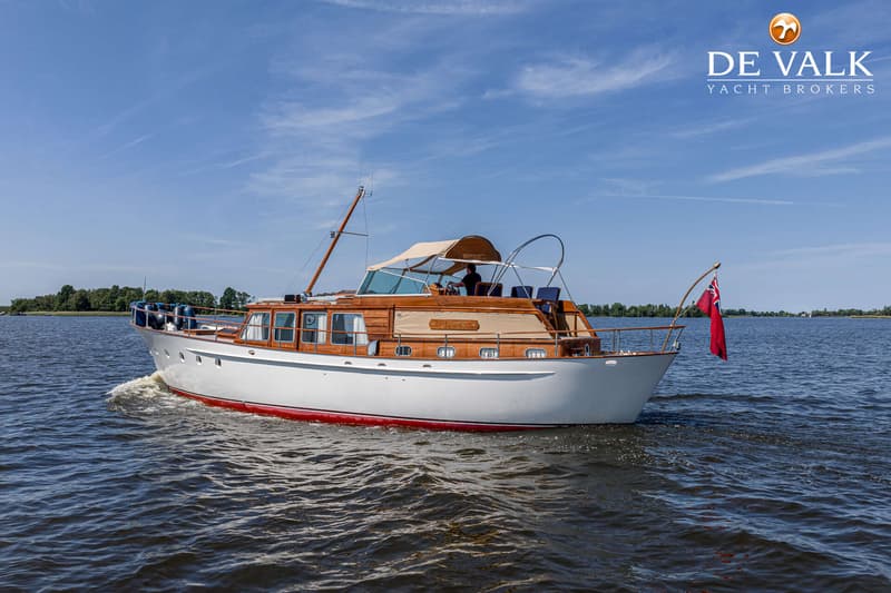 Een witte motorboot met houten opbouw en een rode onderkant vaart op het water onder een blauwe lucht.